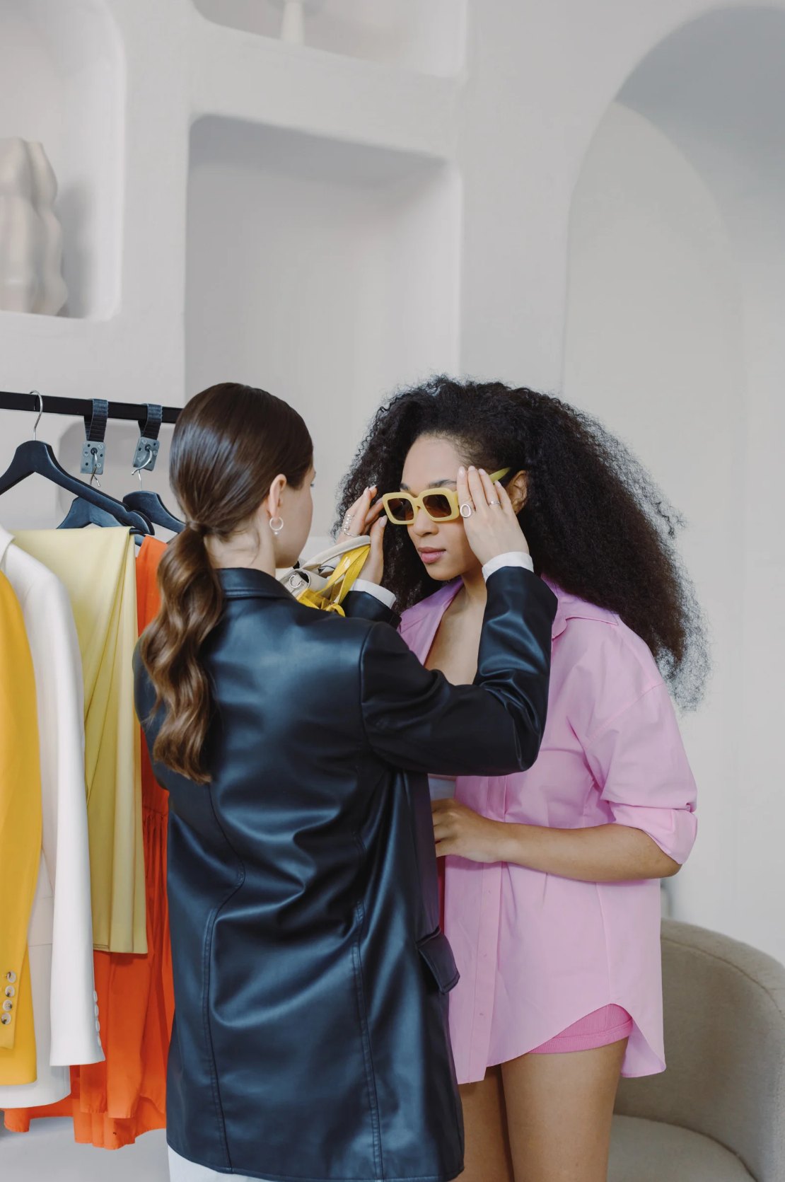 A stylist helping a client try on yellow sunglasses beside a rack of colorful clothing